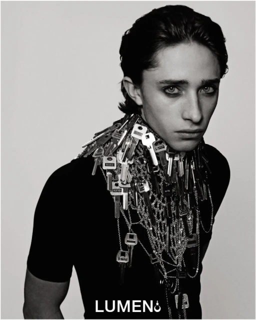 Monochrome portrait of a young adult wearing a black top and an elaborate key necklace against a neutral backdrop.