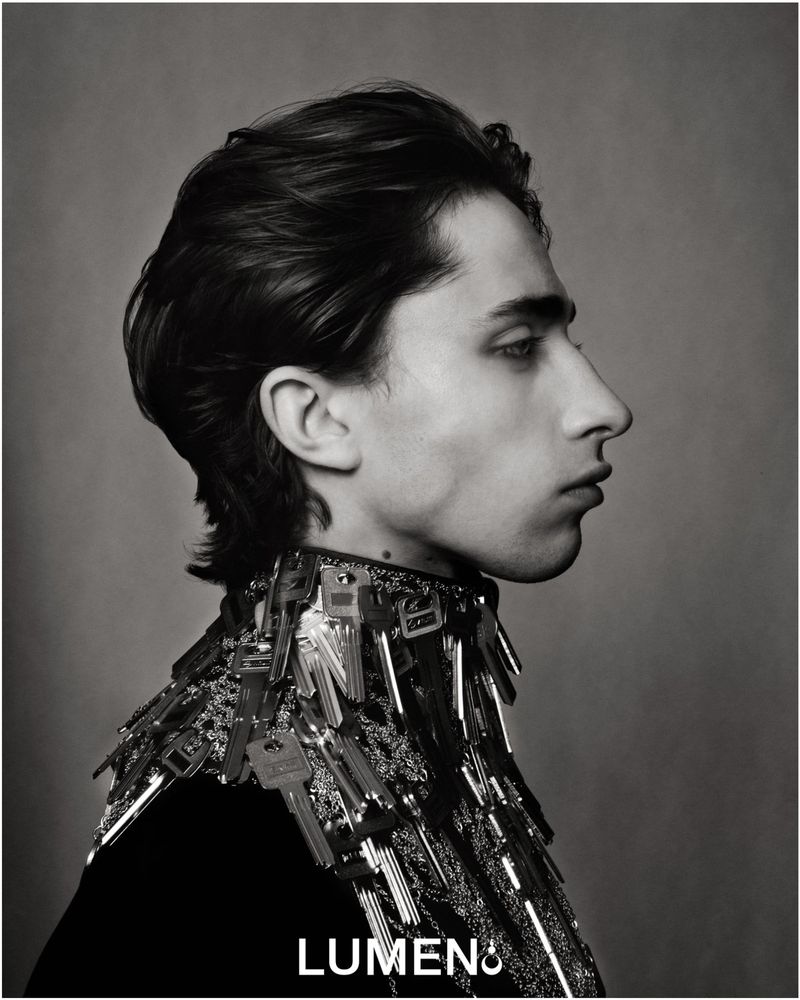 Monochrome portrait of a young adult with dark hair in profile, wearing a key-covered garment, captured in a studio setting.