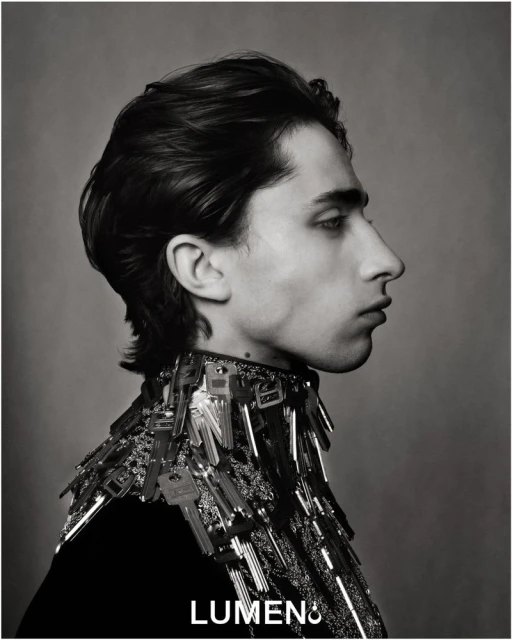 Monochrome portrait of a young adult with dark hair in profile, wearing a key-covered garment, captured in a studio setting.