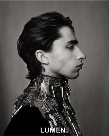 Monochrome portrait of a young adult with dark hair in profile, wearing a key-covered garment, captured in a studio setting.