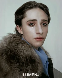 Close-up portrait of Raphael Luce with a tear on his cheek, wearing a fur-collared jacket and blue shirt against a neutral background.