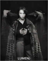 Monochrome portrait of Raphael Luce draped in a houndstooth patchwork cape with translucent arms extended on a dark studio backdrop.