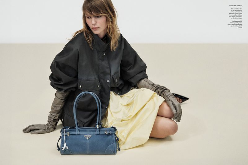 A woman sits on the floor wearing a black jacket, yellow skirt, and grey gloves with a blue Prada bag in the foreground.
