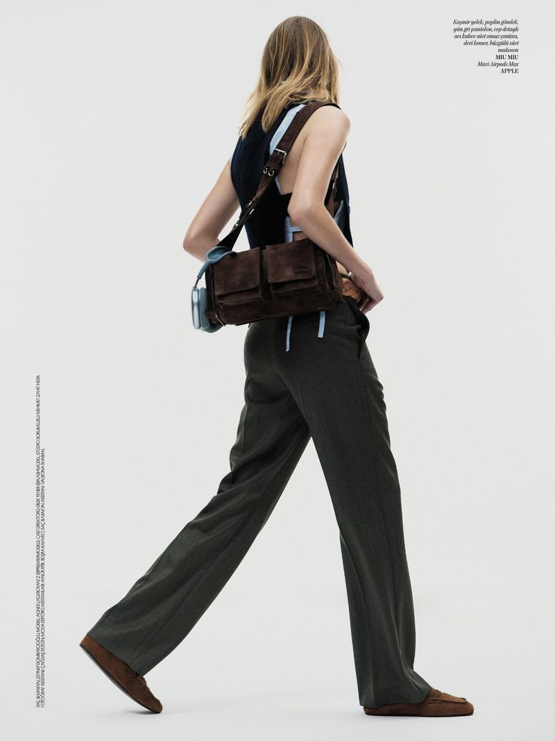 A woman is walking in a sleeveless shirt and trousers, with a brown bag over her shoulder against a plain white background.