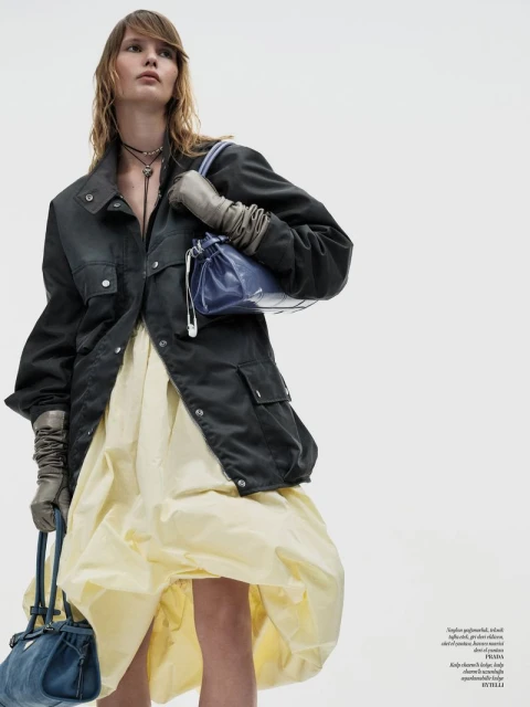 Fashion editorial image of a woman wearing a dark jacket and puffy yellow dress, holding blue purses against a white backdrop.
