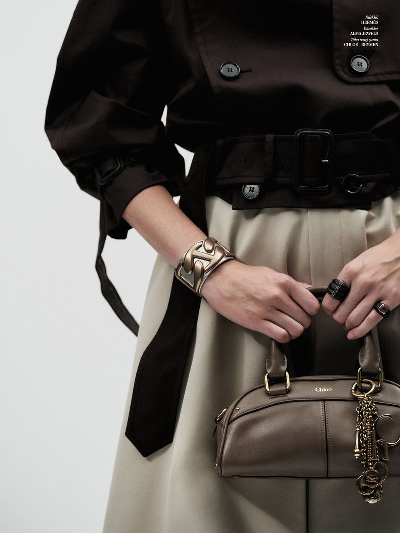 Fashionable adult woman wearing a dark belted jacket and bracelet holds a brown handbag with a key chain in a studio setting.