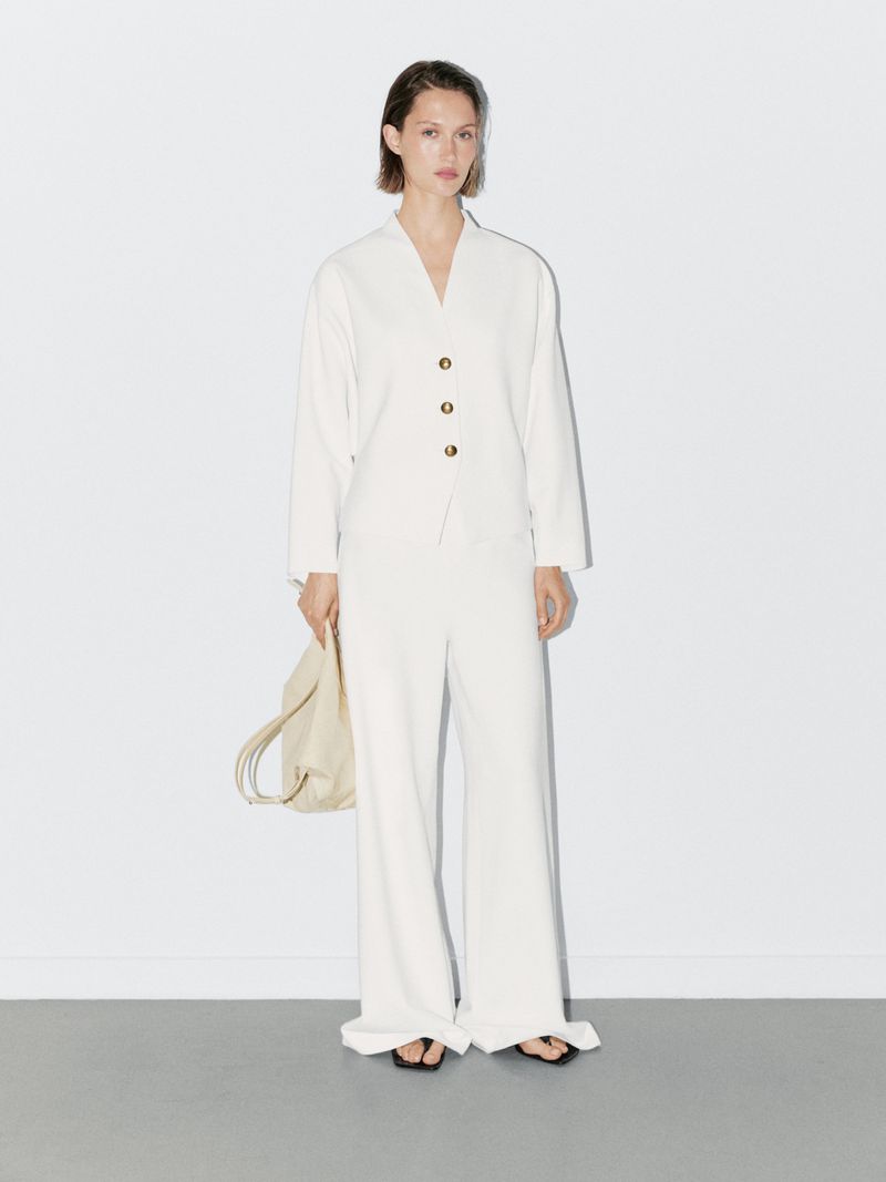 Woman in a light beige suit stands against a white wall, holding a bag. Minimalist fashion shoot with simple styling and neutral colors.