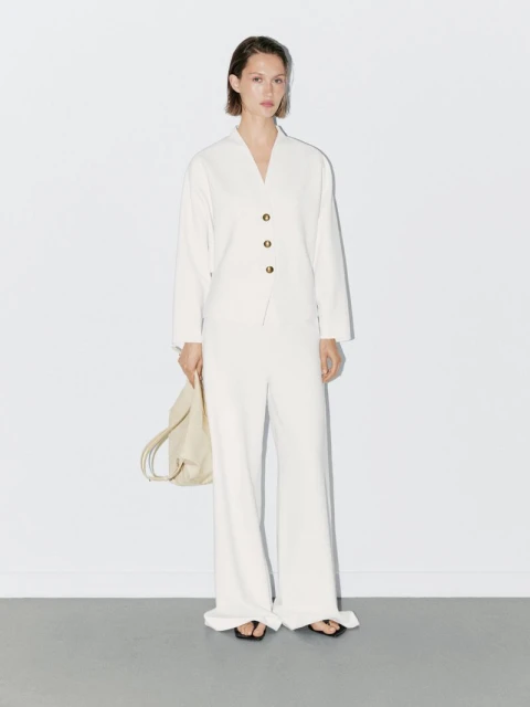 Woman in a light beige suit stands against a white wall, holding a bag. Minimalist fashion shoot with simple styling and neutral colors.