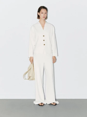 Woman in a light beige suit stands against a white wall, holding a bag. Minimalist fashion shoot with simple styling and neutral colors.