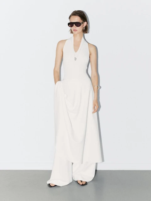 A woman in a white halter dress with wide-leg pants, sunglasses, and jewelry, poses against a bright white background in a studio setting.