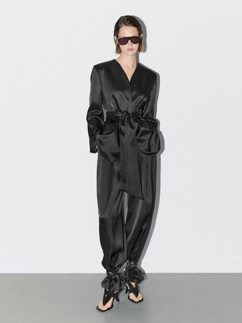 A slender adult female in a black satin wrap jacket, matching pants, and black sandals stands in a brightly lit white photo studio.