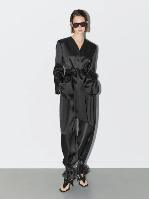 A slender adult female in a black satin wrap jacket, matching pants, and black sandals stands in a brightly lit white photo studio.
