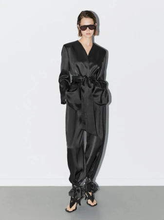 A slender adult female in a black satin wrap jacket, matching pants, and black sandals stands in a brightly lit white photo studio.