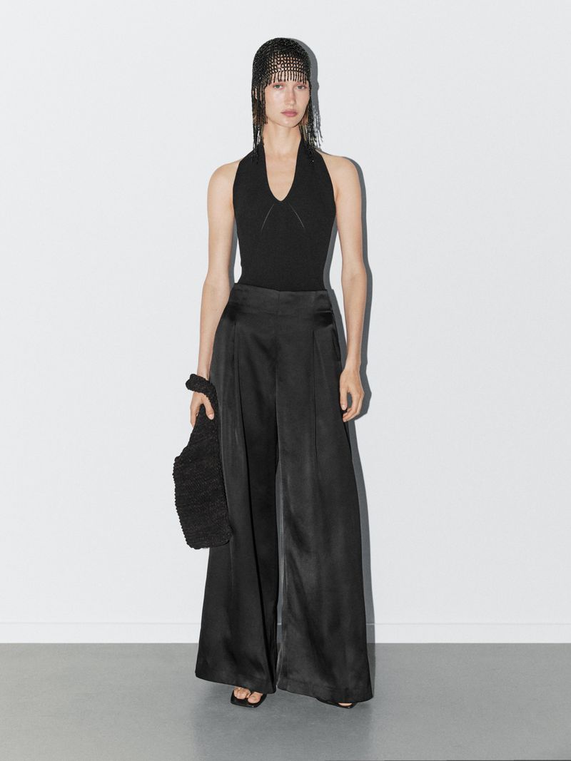 Full-length studio shot of a woman in a black halter top and wide-leg satin pants, holding a furry bag, against a white backdrop.
