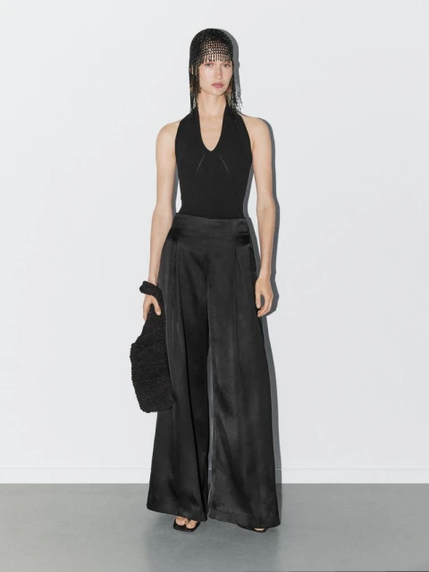 Full-length studio shot of a woman in a black halter top and wide-leg satin pants, holding a furry bag, against a white backdrop.