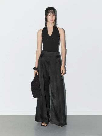 Full-length studio shot of a woman in a black halter top and wide-leg satin pants, holding a furry bag, against a white backdrop.