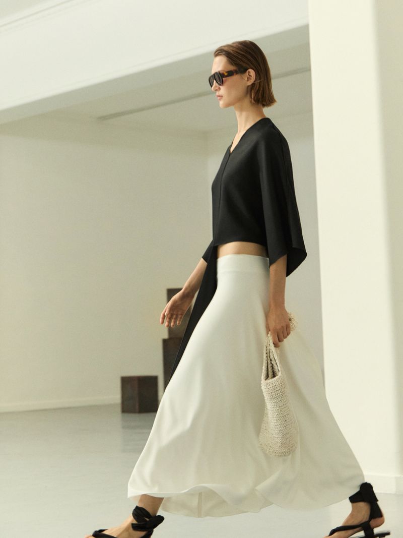 A woman walks in a black top, white skirt, and sunglasses, holding a small white bag, in a minimalist indoor setting with natural light.