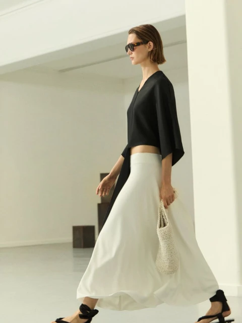 A woman walks in a black top, white skirt, and sunglasses, holding a small white bag, in a minimalist indoor setting with natural light.