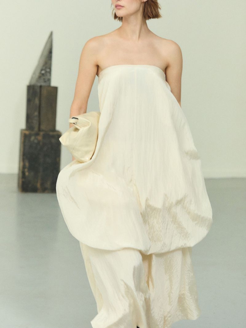 Woman in a floor-length strapless ivory dress poses in front of a sculpture in a bright room during a fashion shoot.
