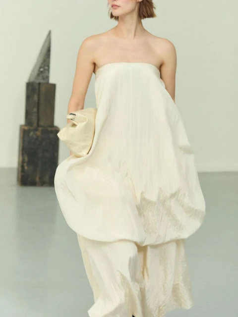 Woman in a floor-length strapless ivory dress poses in front of a sculpture in a bright room during a fashion shoot.