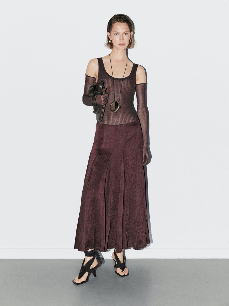 Full-length shot of a slender model posing in a ribbed top, maroon skirt, open-toe heels, and jewelry against a white wall.