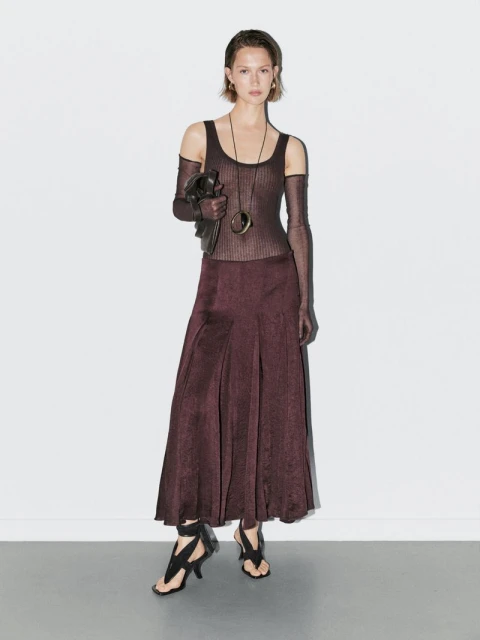 Full-length shot of a slender model posing in a ribbed top, maroon skirt, open-toe heels, and jewelry against a white wall.