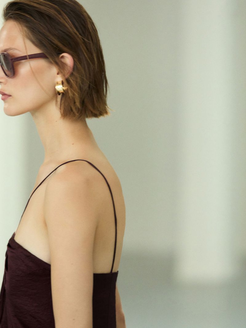 A woman with brown hair and sunglasses is captured in a medium shot, showcasing her minimalist burgundy dress and gold hoop earrings.