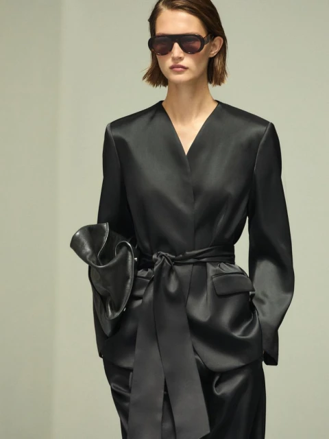 A woman in a black satin suit and sunglasses poses in front of a light gray backdrop. The suit has a large sash and pockets.
