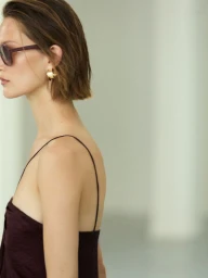 Profile view of a woman wearing sunglasses, hoop earrings, and a dark sleeveless top against a bright minimalist studio background.