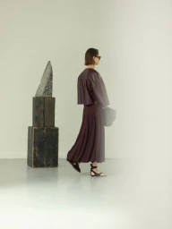 Model in a burgundy jacket and pleated skirt walking against a white seamless background with a sculptural block prop.