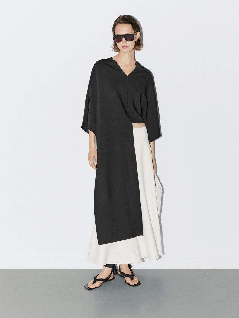 Model wearing dark sunglasses, asymmetric black tunic, and white skirt against a white studio background