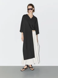 Model wearing dark sunglasses, asymmetric black tunic, and white skirt against a white studio background