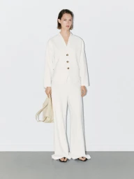 Model wearing a white V-neck buttoned jacket and matching trousers holding a cream handbag against a neutral wall.