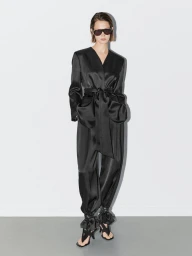 Model in black sunglasses wearing a glossy black belted jacket and trousers against a pale seamless studio background