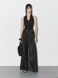 Model in black halter top and wide-leg trousers wearing a mesh headpiece against a plain grey studio backdrop