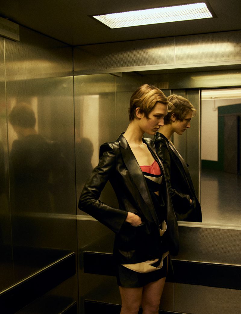 Blonde woman in black leather blazer dress posing in reflective elevator with geometric patterns and sleek metal finish, warm lighting.