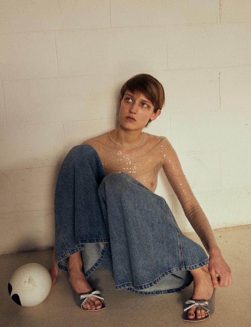 A woman in a sheer top and jeans sits indoors next to a white ball, looking off to the side in a fashion shot with soft lighting.