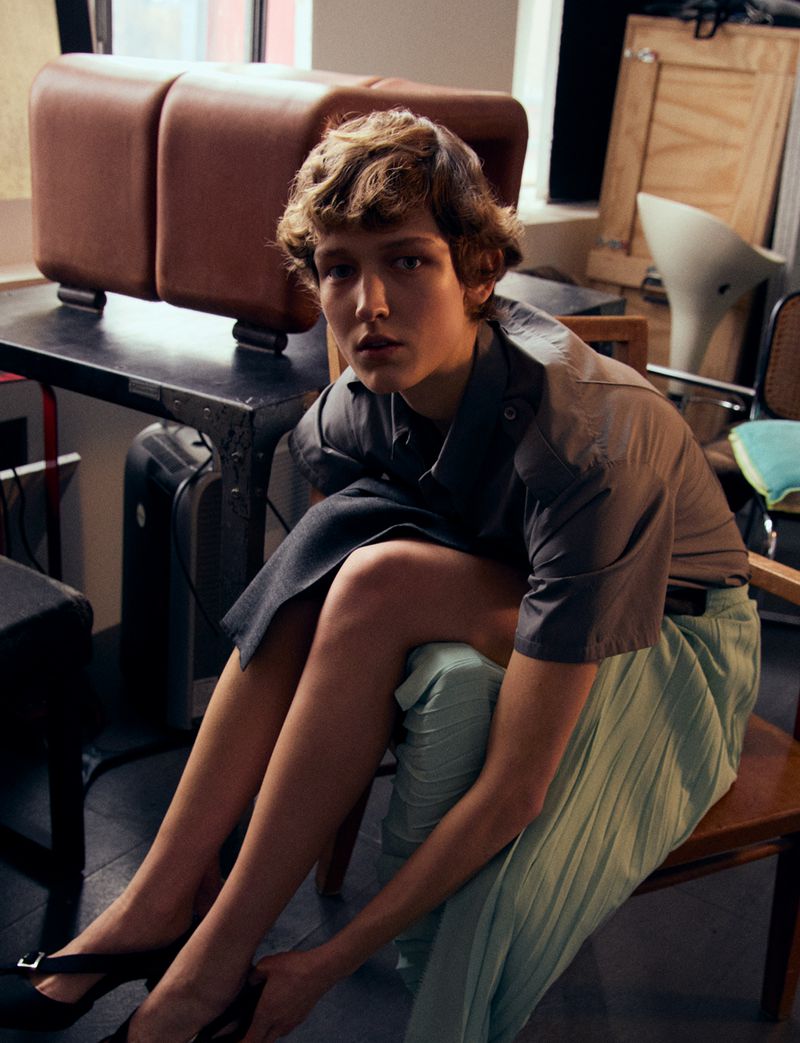 Young woman in casual outfit sits on a chair indoors, tying the strap of her black high-heeled shoe, with assorted furniture pieces nearby.