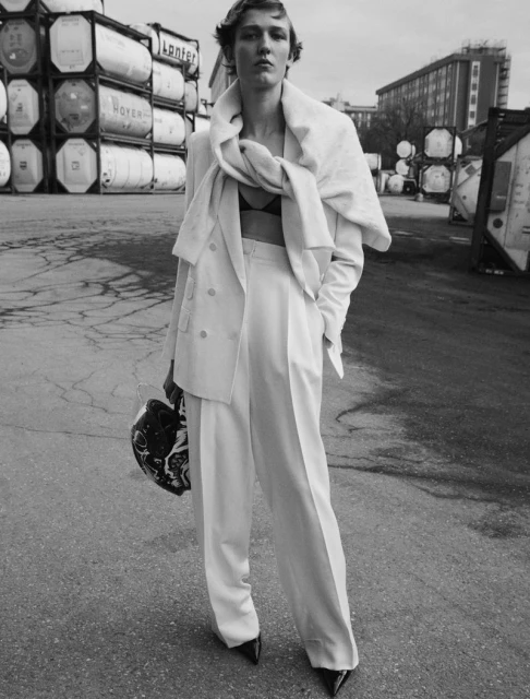 Monochrome portrait of a woman in a white suit with a helmet, standing in an industrial area with shipping containers.
