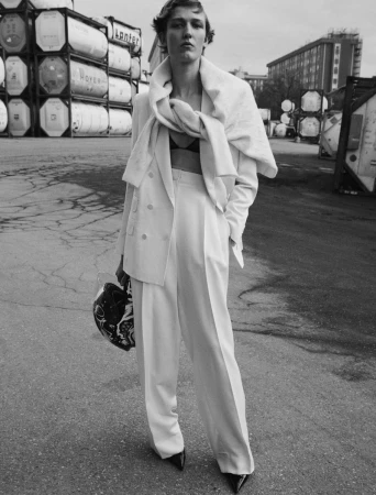 Monochrome portrait of a woman in a white suit with a helmet, standing in an industrial area with shipping containers.