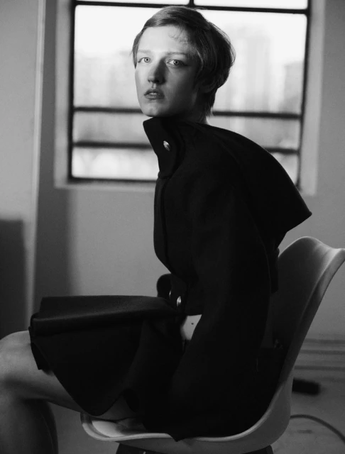 Black and white portrait of a young adult female with a short haircut, posing while sitting on a chair near a window.