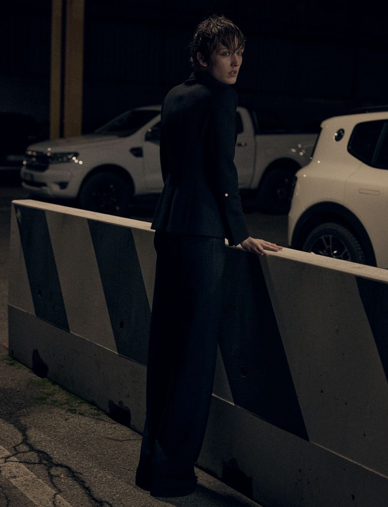 A female fashion model in a dark suit poses in an urban environment at night, leaning against a striped barrier with cars in the background.