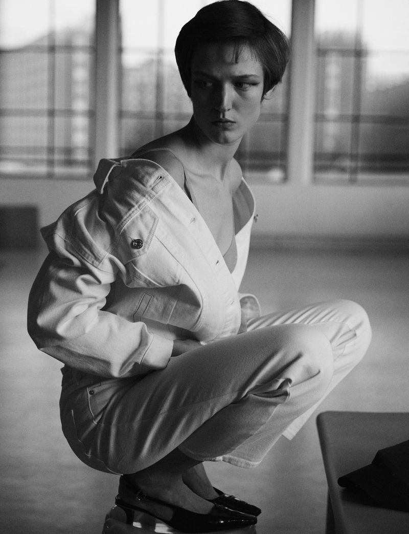 Monochrome portrait of a crouched woman in a white denim jacket, looking off-camera in a bright, indoor setting with architectural details.