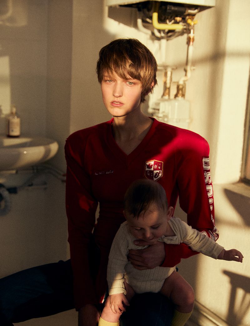 Indoor shot of a light-skinned adult holding a fair-skinned baby in a red V-neck shirt, with a sink, radiator, and window in the background.