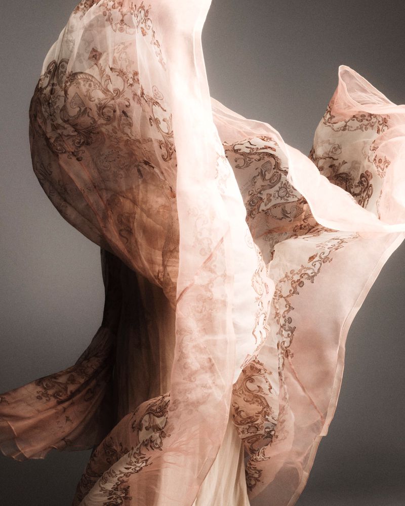 Flowing sheer fabric with an ornate print, gracefully suspended in mid-air against a soft gray backdrop creating an ethereal fashion moment.