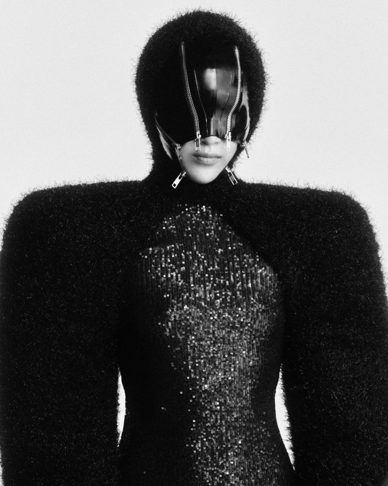 Person wearing a black fuzzy garment and zippered mask poses against a white background in a studio shot, fashion forward and unusual.