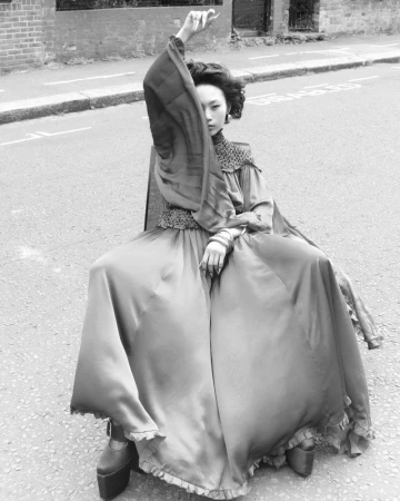 A fashion shot of a woman in a long dress sitting on a chair on a street with buildings in the background, presented in black and white.