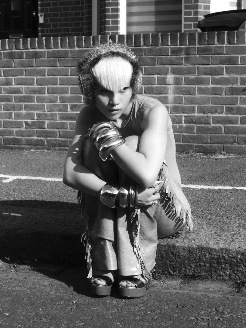 Model Wawa Li crouching on a street curb wearing fringed trousers, platform sandals, and chunky metallic arm cuffs in black and white.