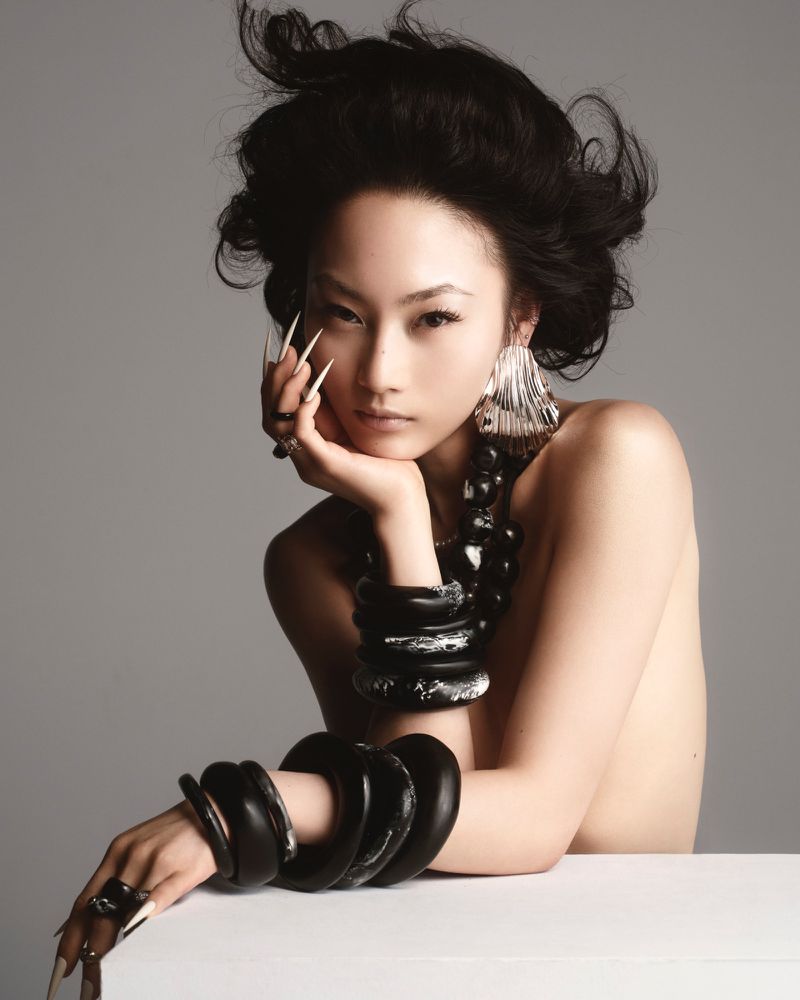 Glamour shot of an Asian woman with long nails, statement earrings, and jewelry posing in a photo studio against a neutral background.