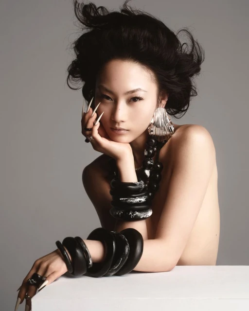 Glamour shot of an Asian woman with long nails, statement earrings, and jewelry posing in a photo studio against a neutral background.